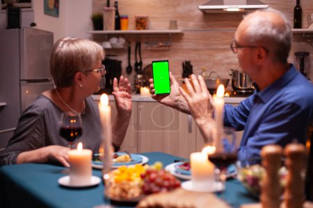 Téléchargez les photos : Homme et femme âgés saluant au téléphone avec écran vert. Personnes âgées regardant modèle de maquette chroma clé isolée écran de téléphone intelligent en utilisant la technologie Internet assis à la table dans la cuisine. - en image libre de droit