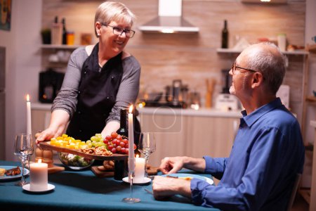 Photo for Senior woman serving husband with grapes and nuts as appetizer for romantic dinner. Elderly old couple talking, sitting at the table in kitchen, enjoying the meal, celebrating their anniversary . - Royalty Free Image