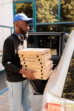 Téléchargez les photos : Livreur portant pizza au bureau en voiture vue de côté, homme afro-américain tenant des boîtes pile. Déjeuner service de livraison employé livrant fastfood, debout près de l'immeuble de l'entreprise à l'extérieur - en image libre de droit