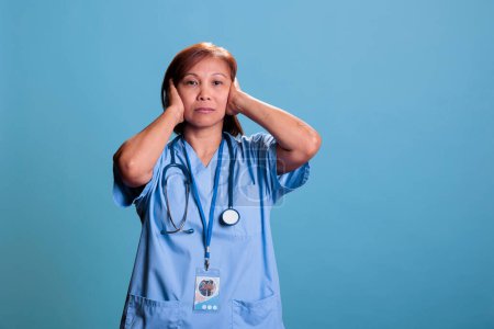 Photo for Asian quiet nurse with stethoscope covering ears having serious expression during checkup visit consultation. Medical assistant doing three wise monkeys symbol. Hear no evil concept. - Royalty Free Image