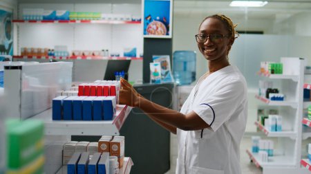 Foto de Una farmacéutica sonriente analizando cajas de medicamentos en los estantes de la farmacia, mirando las vitaminas y los frascos de pastillas para ayudar a los clientes. Empleado que trabaja en farmacia con productos farmacéuticos. - Imagen libre de derechos