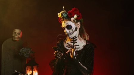 Junge gruselige Modell mit Handy, um Internet-Website über schwarzen Hintergrund surfen. Santa muerte tote Frau, die Horrorgöttin des Todes auf mexikanischen Feiertagen darstellt. Handschuss.