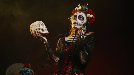 Photo for Mexican horror goddess on phone call holding skull, talking on smartphone line and acting creepy in studio. Answering telephone for remote chat, wearing holy mexican entity costume. Handheld shot. - Royalty Free Image