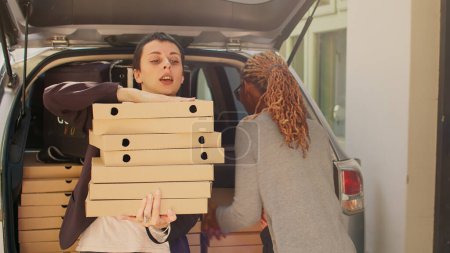 Photo for Food delivery employee delivering big order of pizza boxes to customer, giving meal packages of food from vehicle trunk to people. Pizzeria courier taking fastfood stacks for delivery service client. - Royalty Free Image