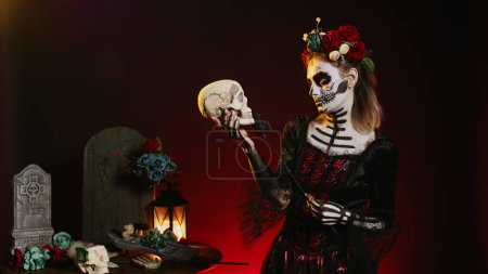 Foto de Hermosa mujer vestida con maquillaje tradicional para celebrar la fiesta mexicana, sosteniendo el cráneo en el estudio. Pareciendo santa muerte o diosa de la muerte en la celebración del santo dios de los muertos. - Imagen libre de derechos
