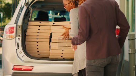 Téléchargez les photos : Employé de Pizzeria donnant des piles de boîtes de pizza à divers clients, prendre des paquets de repas hors du coffre du véhicule. Courrier de livraison de nourriture livrant des piles de fastfood aux clients qui attendent près de la porte d'entrée. - en image libre de droit