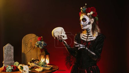 Foto de Mujer muerta espeluznante sosteniendo el cráneo sagrado en el estudio, actuando de miedo y horror para celebrar el día de Halloween mexicano. Diosa coqueta vistiendo disfraz de festival con arte corporal, pareciendo dama de la muerte. - Imagen libre de derechos