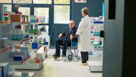 Téléchargez les photos : Pharmacien donnant bouteille de pilules de cardiologie à l'utilisateur de fauteuil roulant aîné, homme souffrant d'un handicap chronique demandant de l'aide. Client ayant une déficience recevant un traitement pharmaceutique en pharmacie. - en image libre de droit