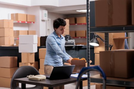 Photo for Supervisor putting trendy blouse in cardboard boxes preparing customers orders for delivery, checking shipping detalies on laptop computer in storehouse, Worker analyzing transportation logistics - Royalty Free Image