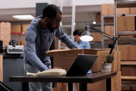 Photo for Worker putting client order in cardboard box wrapping with bubble wrap for protection, checking shipping details on laptop computer before delivery package. Employee working in distribution center - Royalty Free Image