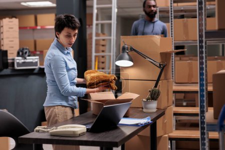 Photo for Employee putting trendy clothes in cardboard boxes preparing clients orders for delivery, checking shipping detalies on laptop computer in storehouse, Worker analyzing transportation logistics - Royalty Free Image