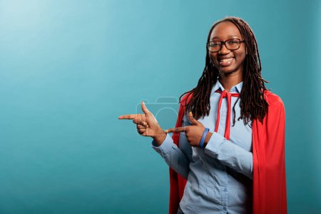 Foto de Brave and proud justice defender wearing mighty hero cape while pointing fingers to left on blue background. Positive and happy young adult justice defender person smiling heartily at camera. - Imagen libre de derechos