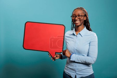 Foto de African american confident and smiling heartily woman holding red cardboard speech bubble on blue background. Joyful young adult woman holding blank template announcement placard. - Imagen libre de derechos