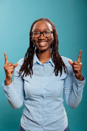 Téléchargez les photos : Excited happy beautiful woman smiling heartily while pointing fingers up on blue background. Confident and positive young adult person pointing hands up while looking at camera. - en image libre de droit