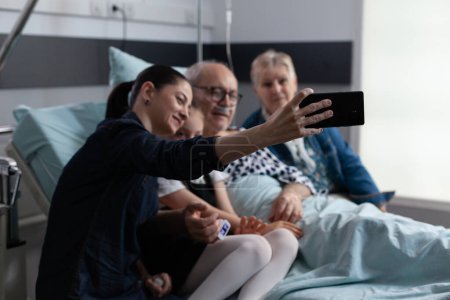 Photo for Accompanying hospitalized elderly man for routine checkups capturing cell phone snapshots at geriatric medical tower. Young woman taking selfie with sick father in clinic rest room. - Royalty Free Image