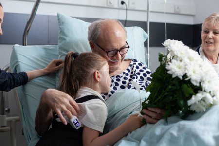 Foto de Abuelo abrazando a su nieta en la habitación del hospital. Familiares ancianos enfermos mostrando afecto antes de salir de la clínica médica. Paciente postrado en cama recibiendo regalo de familiares. - Imagen libre de derechos