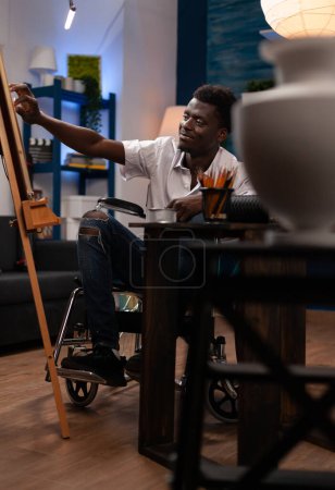 Téléchargez les photos : Artiste vivant avec un handicap faire un vase dessin sur toile. Heureux étudiant en art afro-américain créant des œuvres d'art avec des crayons de couleur au home studio. Utilisateur de fauteuil roulant l'apprentissage de nouvelles compétences artistiques. - en image libre de droit