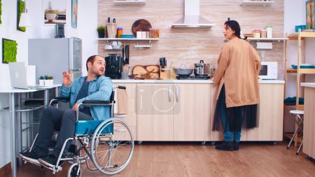 Téléchargez les photos : Heureux homme handicapé en fauteuil roulant en utilisant un ordinateur portable dans la cuisine. Homme parlant en vidéoconférence. Entreprise homme avec paralysie handicap handicap difficultés à travailler après un accident ayant - en image libre de droit