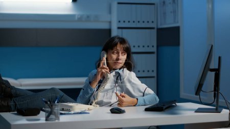 Photo for Relaxed doctor sitting with feet on desk table while talking at phone whith remote friend during night shift in hospital office. Physician woman working at medical report analyzing expertise. - Royalty Free Image