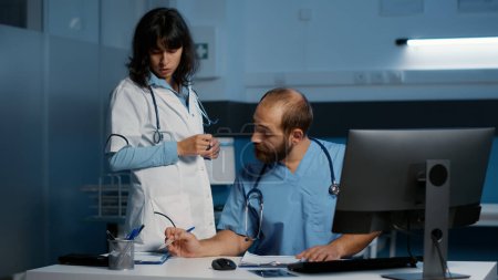 Photo for Medical team analyzing patient disease report on computer while working over hours in hospital office. Doctor and assistant planning health care treatment to help cure illness. Medicine service - Royalty Free Image