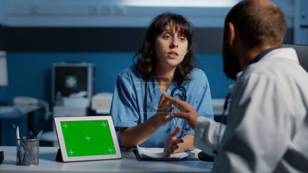 Téléchargez les photos : Assistant discutant de l'expertise du patient avec le médecin tout en analysant l'ordinateur tablette avec le modèle d'écran vert lors de la visite de contrôle. Personnel clinique travaillant tard dans la nuit au bureau de l'hôpital - en image libre de droit