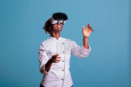 Téléchargez les photos : Chef professionnelle en uniforme jouant à des jeux de réalité virtuelle avec des lunettes VR, explorant les simulations. Un cuisinier afro-américain bouge les bras en touchant des objets dans le cyberespace. - en image libre de droit