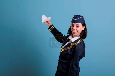 Foto de Asistente de vuelo sosteniendo avión de papel mientras mira a la cámara, azafata jugando con el juguete del avión. Azafata de aire mirando el modelo de jet comercial, con uniforme de aviación, retrato de vista frontal - Imagen libre de derechos