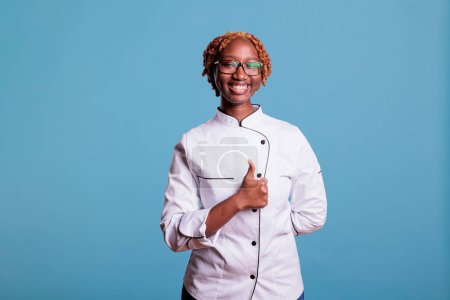 Téléchargez les photos : Afro-américain cuisine professionnelle souriant, avec un geste de main affirmatif. Femme cuisinière aux cheveux bouclés et riante, en accord avec ses pairs. heureux afro restaurant travailleur aimer. - en image libre de droit