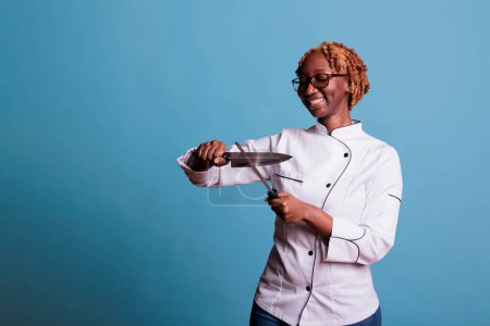 Photo for African american female chef in uniform sharpening professional knife blade before beginning her work shift. Woman preparing kitchenware for cooking, holding sharp culinary object in hands. - Royalty Free Image