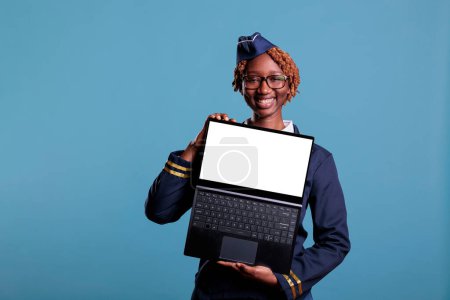Photo for Portrait of stewardess holding laptop with white screen with advertising space looking with a smile at the camera. Female flight attendant with notebook in studio shot against blue background. - Royalty Free Image