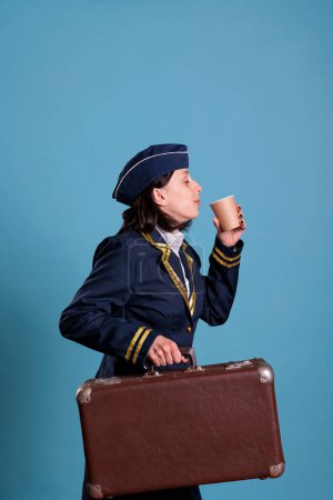 Photo for Flight attendant in professional airline uniform carrying suitcase, drinking coffee to go. Stewardess with luggage in airport, air hostess with baggage holding tea paper cup, side view - Royalty Free Image