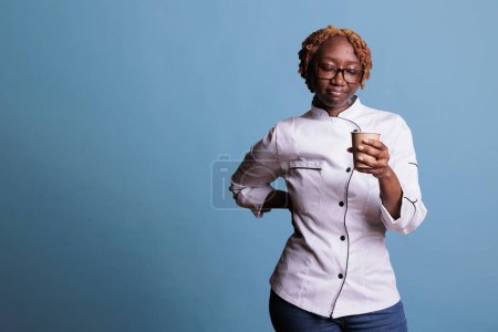 Téléchargez les photos : Cuisinier afro-américain onéreux avec une expression triste en regardant la tasse de café jetable. Femme de cuisine professionnelle portant l'uniforme regardant déçu par les mauvaises nouvelles dans une journée de travail. - en image libre de droit