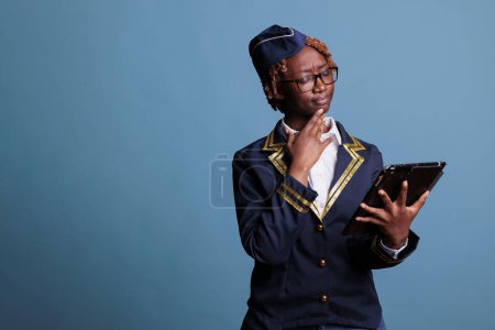 Téléchargez les photos : Hôtesse afro-américaine porte uniforme de la compagnie aérienne regardant attentivement l'écran de la tablette numérique. L'agente de bord vérifiant le nouvel horaire de travail semble confondue avec les changements. - en image libre de droit