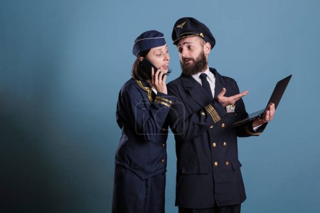 Photo for Smiling aviator using laptop while stewardess talking on smartphone. Airlane captain in professional flight uniform holding portable computer, air hostess chatting on mobile phone - Royalty Free Image