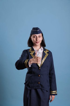 Photo for Smiling flight attendant showing thumbs up gesture, aviation academy stewardess doing approval sign. Young attractive air hostess in professional uniform approving, studio shot - Royalty Free Image