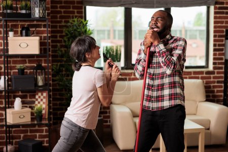 Photo for Multiracial couple singing at home using broom and tv remote control as microphone after finishing spring cleaning. Woman and African American man having fun with household chores. - Royalty Free Image