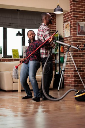 Photo for African american family doing housework in urban apartment, playing musician with mop and vacuum cleaner. Enthusiastic married couple feeling joy while doing household chores. - Royalty Free Image