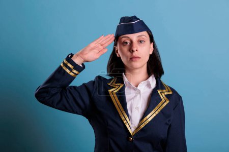 Photo for Young serious flight attendant saluting, wearing uniform, standing, looking at camera. Confident plane stewardess portrait, serious air hostess with hand near head front view, studio medium shot - Royalty Free Image