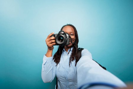 Photo for Creative amateur photographer taking pictures with modern camera for social media. Young adult african american woman pointing DSLR device to camera while taking a photo on blue background. - Royalty Free Image