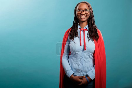 Foto de Positive smiling heartily mighty and brave justice defender standing on blue background while looking at camera. Young adult confident and strong superhero woman wearing hero cloak. Studio shot - Imagen libre de derechos