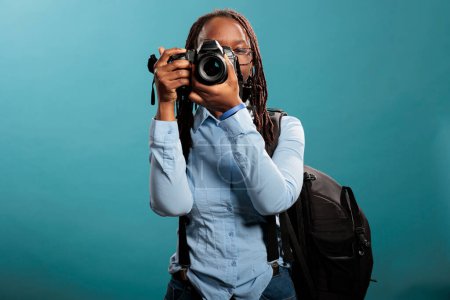 Foto de Portrait of professional photographer with photography modern device taking pictures while standing on blue background. Young adult woman with DSLR camera shooting photos. Studio shot - Imagen libre de derechos