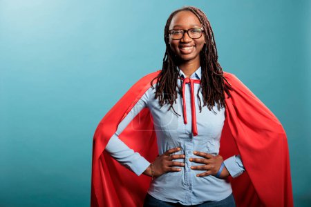 Photo for Proud and happy young superhero woman posing brave for camera while standing on blue background. Strong and selfless justice defender wearing mighty hero red cloak while standing with hands on hip. - Royalty Free Image