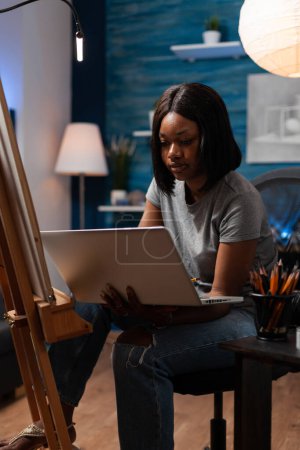 Foto de Mujer afroamericana con intereses artísticos usando laptop para tomar clases de arte a través de videoconferencias desde un estudio creativo. Dibujo estudiante buscando técnicas de dibujo en Internet para hacer obras de arte. - Imagen libre de derechos
