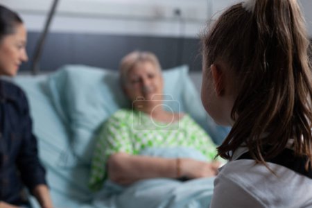 Foto de Mujer mayor fuera de foco, acostada en la cama del hospital charlando con una niña no identificada. Familia visitando a paciente anciana geriátrica en recuperación en sala de clínica médica. - Imagen libre de derechos