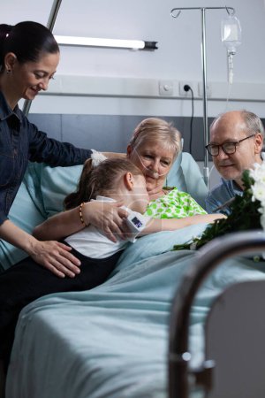 Elderly woman suffering illness lying in hospital observation room bed, hugging young granddaughter during relatives visit. Family giving flowers bouquet to sick grandmother at medical clinic.