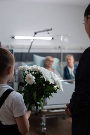 Foto de Pareja mayor visitada por su hija, nieta en la sala de descanso de la clínica geriátrica. Niña llevando flores regalo para recuperar abuelo acostado en la cama del hospital. - Imagen libre de derechos