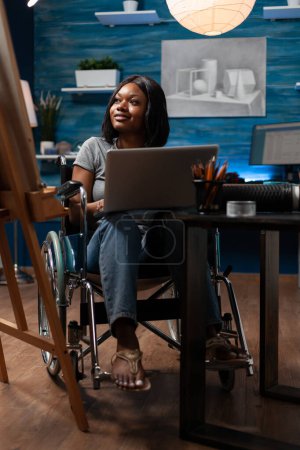 Photo for Female sketch artist wheelchair user using laptop computer to take drawing classes remotely at home made studio. African american looking for inspiration online to create artwork. - Royalty Free Image