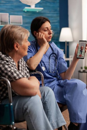 Téléchargez les photos : Travailleur soignant aidant une femme retraitée handicapée ont un appel vidéo sur smartphone avec un médecin. Assistante sociale travaillant avec des personnes handicapées dans une maison de soins infirmiers. - en image libre de droit