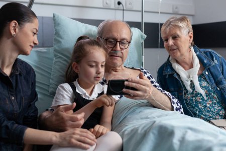 Photo for Little girl showing cell phone videos to grandfather hospitalized in geriatric clinic. Elderly man looking family photos at hospital room. Sick man relatives showing pictures on smartphone. - Royalty Free Image