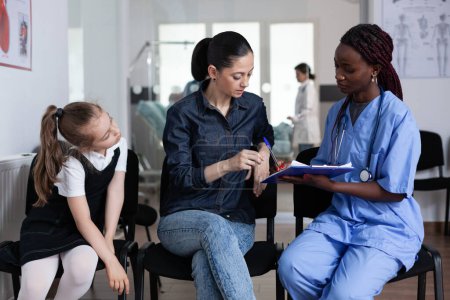 Photo for Young woman signing little girl discharge papers in children hospital waiting room. Mother filling out daughter file with african american pediatrician at medical clinic lobby. - Royalty Free Image
